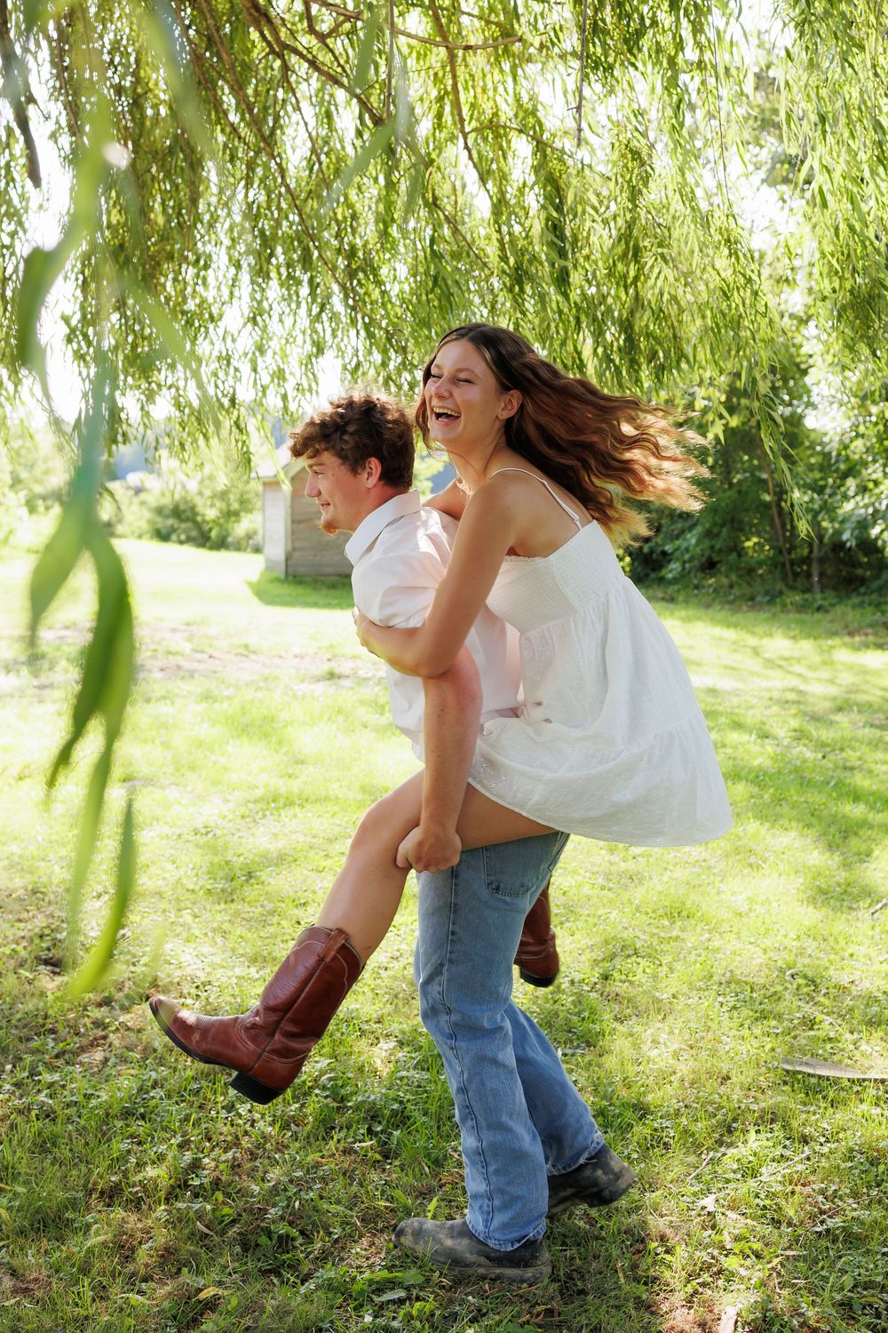 A playful couple dances and spins together beneath drooping willow branches in a sunlit garden.
