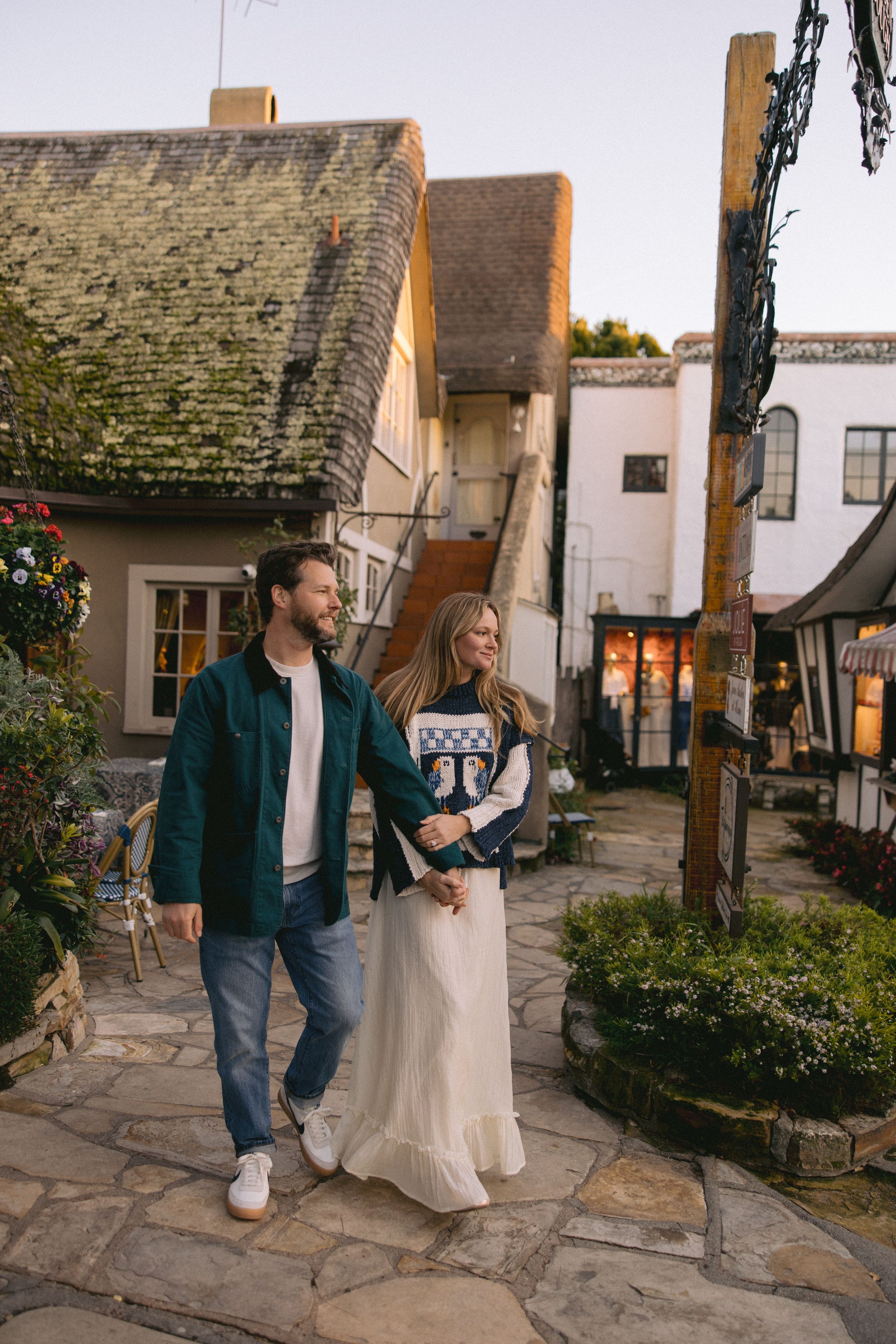 A couple strolls along a stone path by rustic old buildings with moss-covered roofs in a charming village setting.