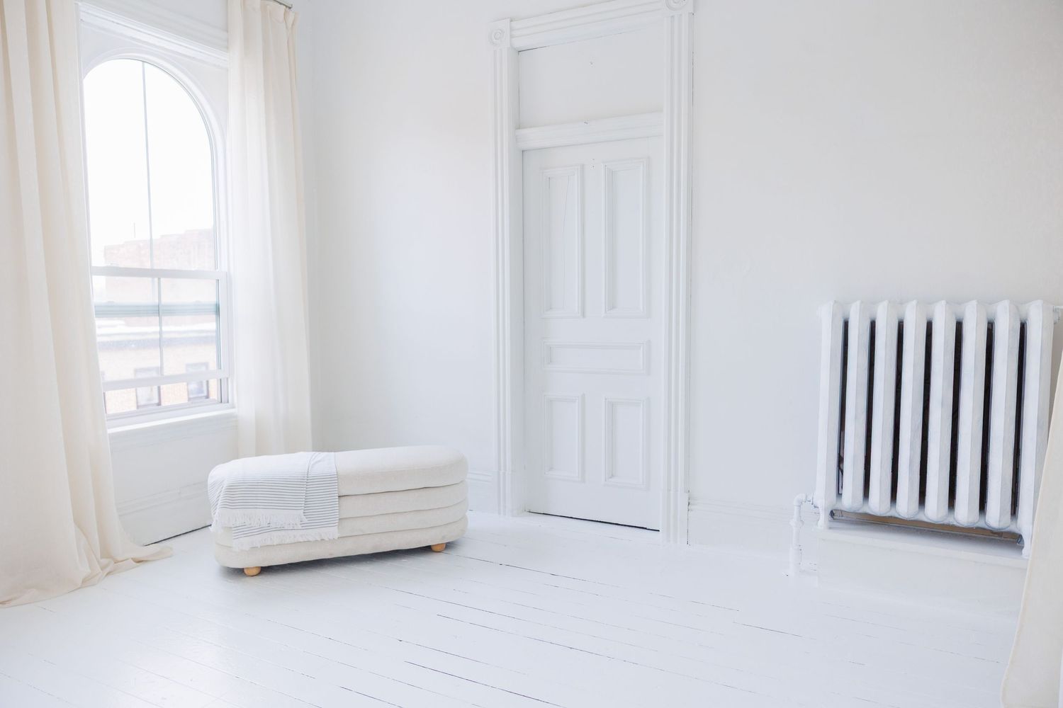 Minimalist white room interior with arched window, radiator, and ottoman in bright natural light.