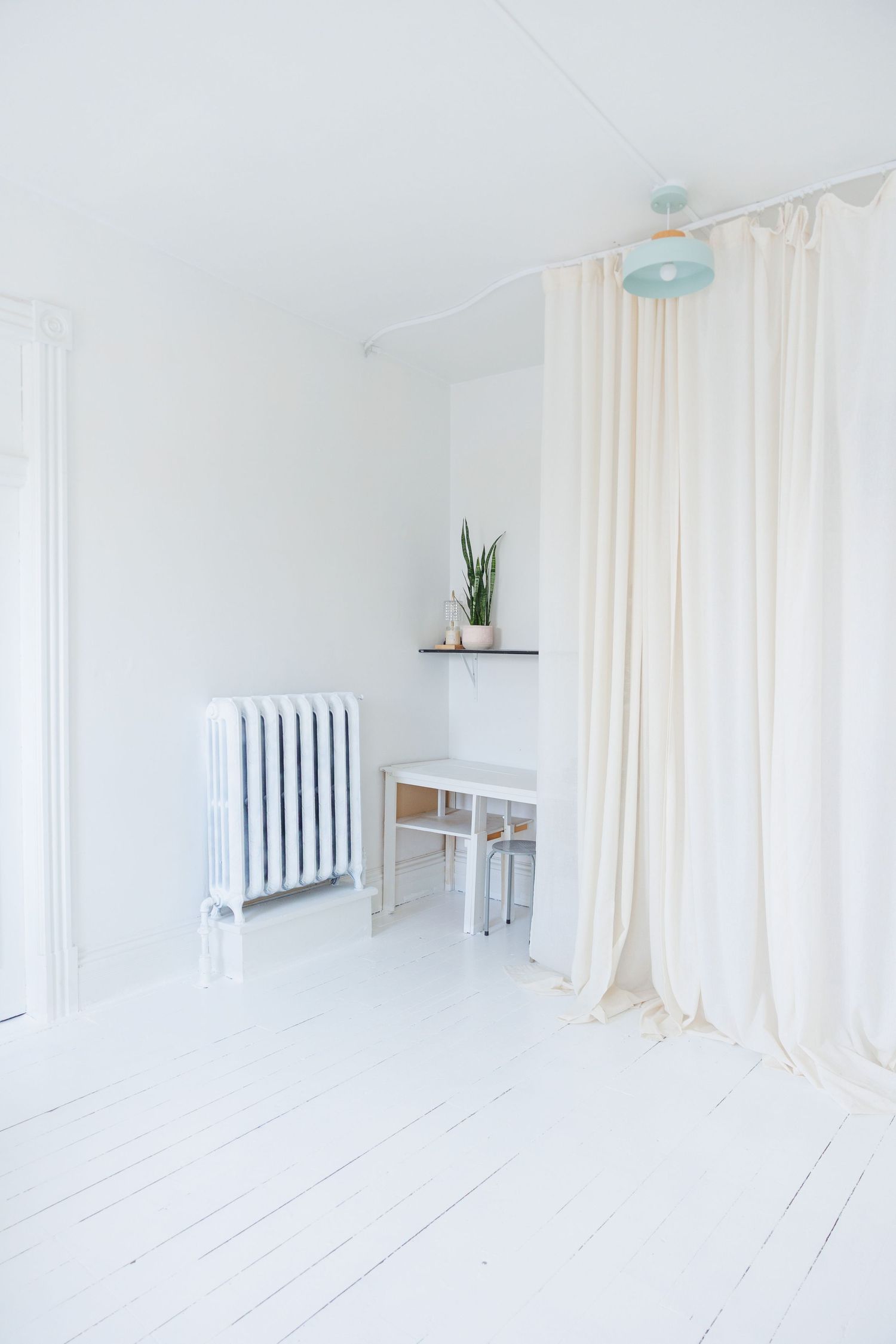 Minimalist white interior room with radiator, flowing curtains, and painted wooden floors in bright natural light.