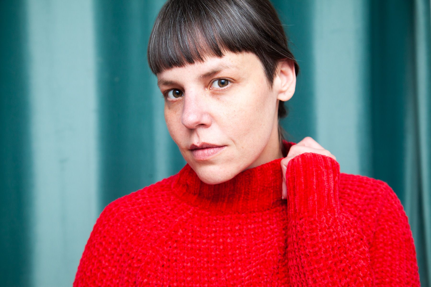Against a teal background, a figure in a bright red knitted sweater gazes directly at the camera.