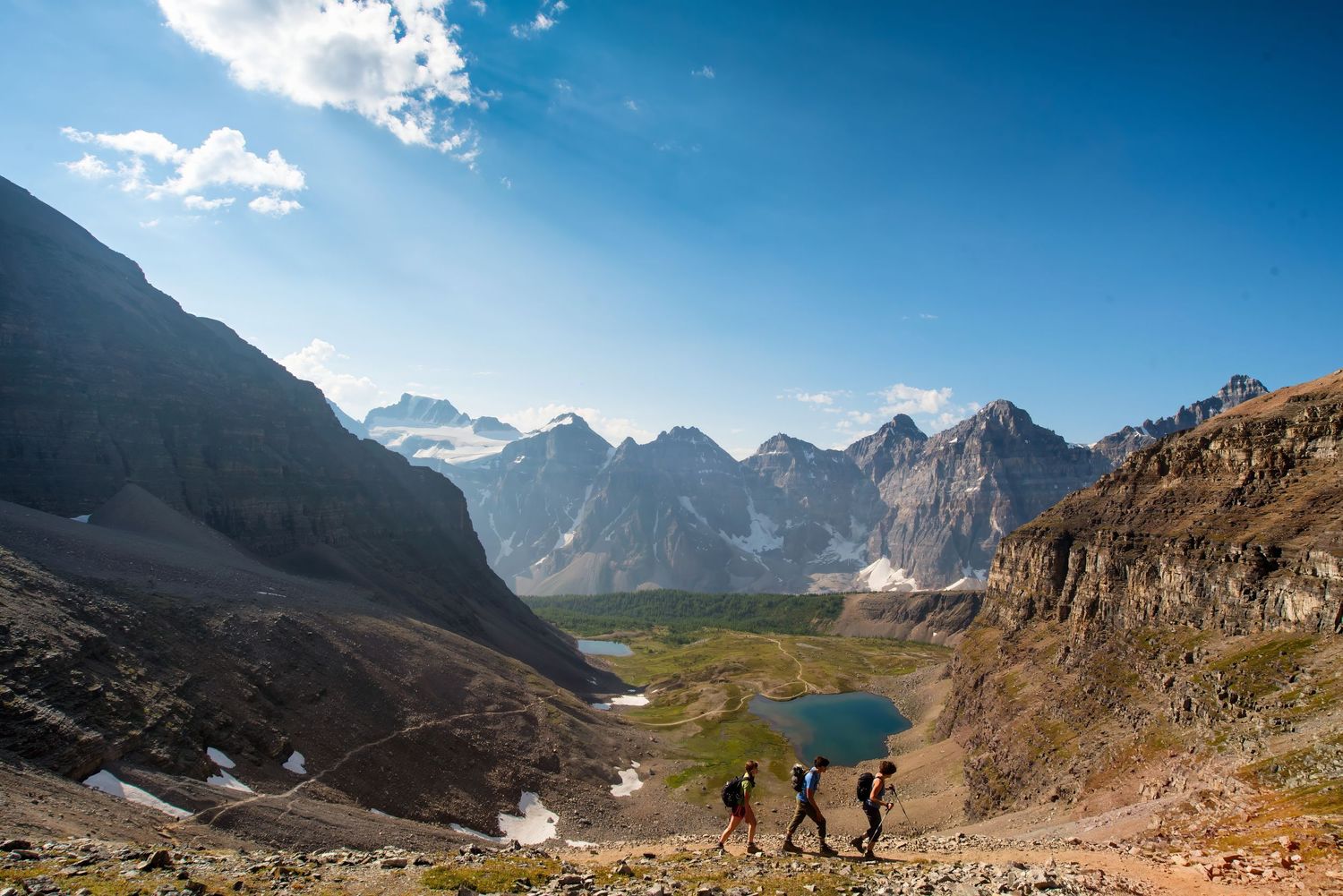 Two hikers descending a rocky mountain trail overlooking a turquoise alpine lake and jagged peaks in the distance.