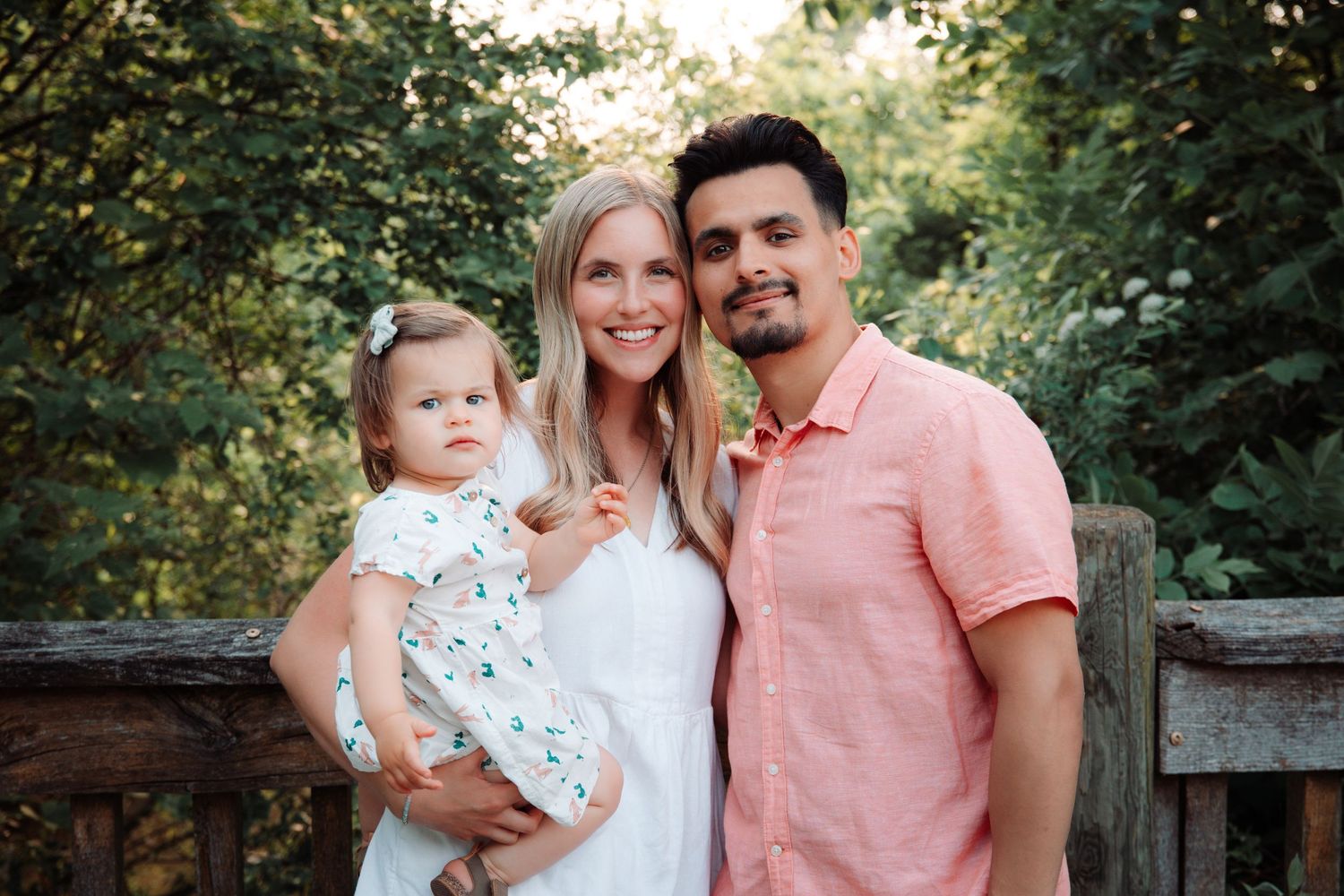 A family of three poses together on a wooden deck during golden hour in a garden setting.