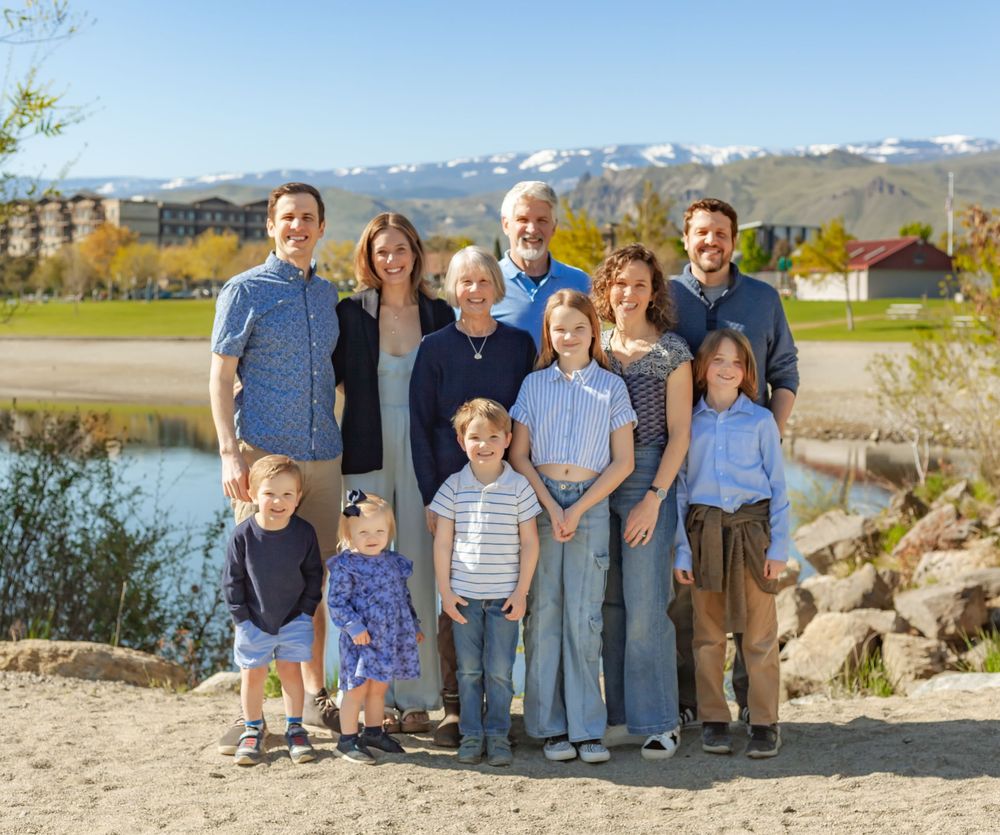 A family group poses outdoors near a lake with snow-capped mountains and fall foliage in the background.
