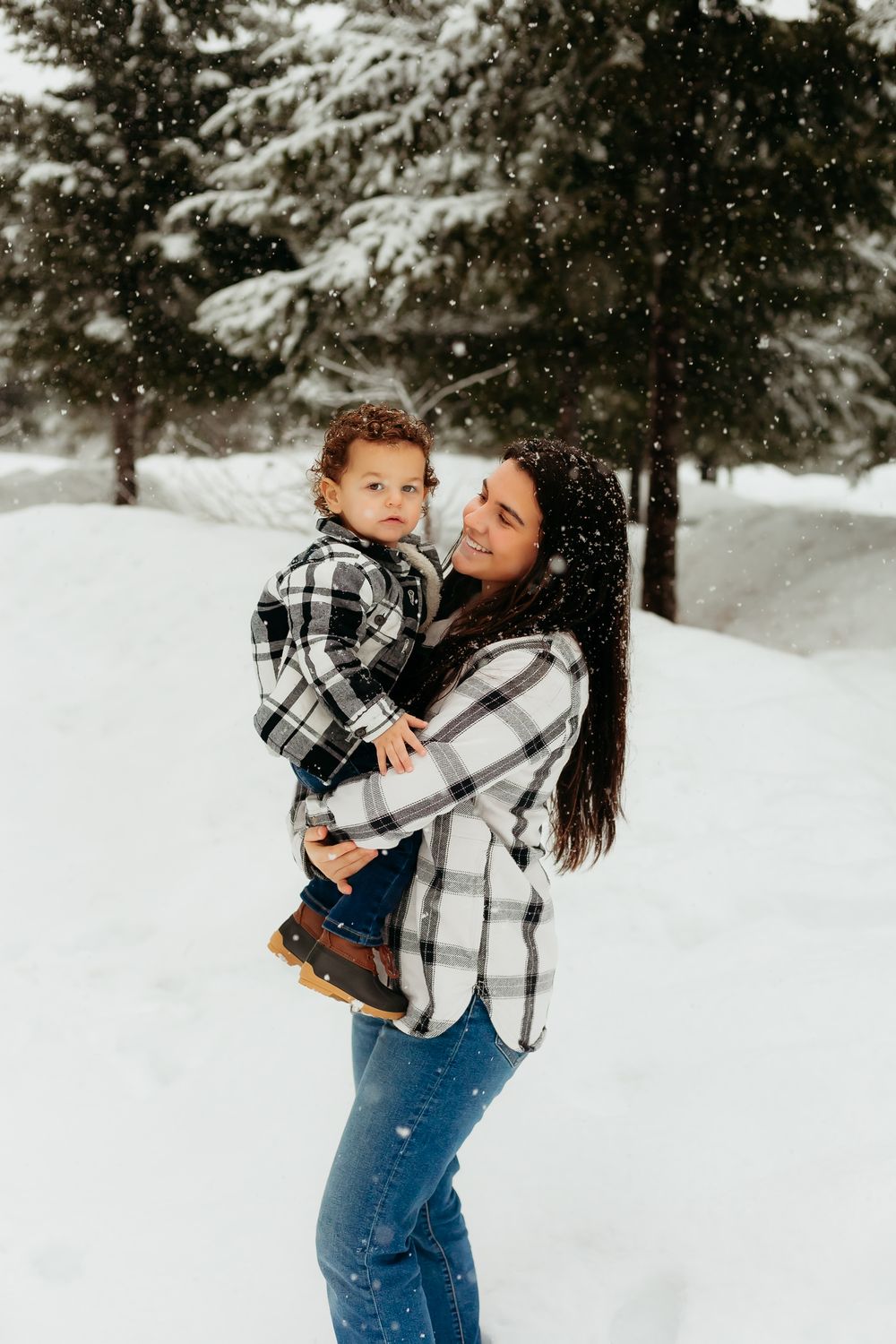 Parent and child in matching plaid flannel shirts embrace during a snowy winter photo session in the forest.