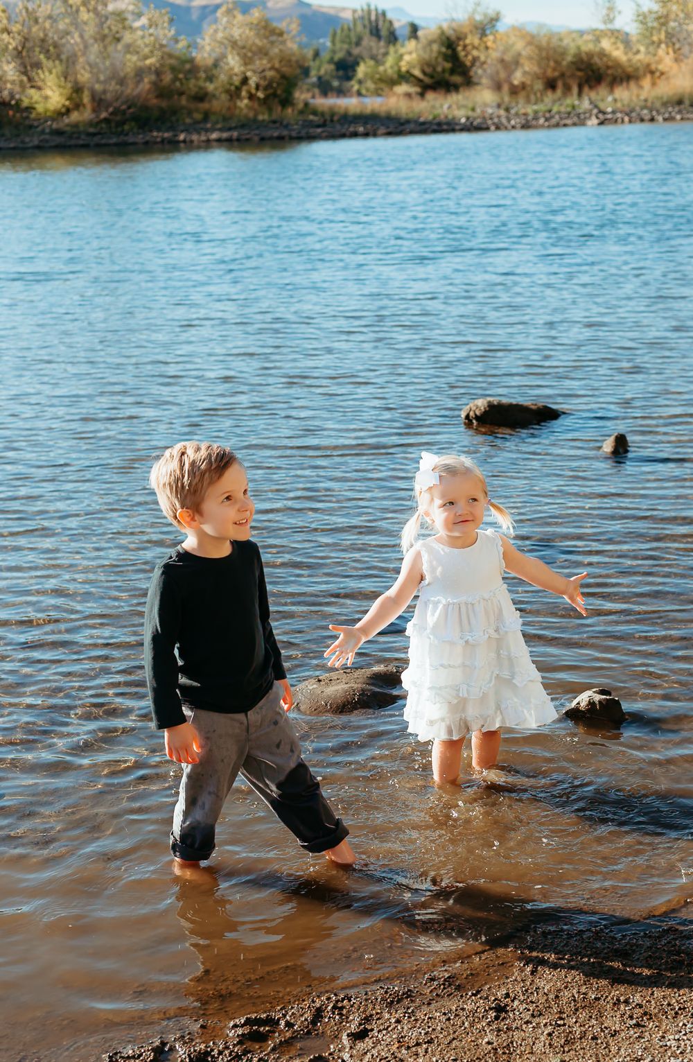 Two children wade in shallow water at a scenic lake surrounded by mountains and autumn trees on a sunny day.