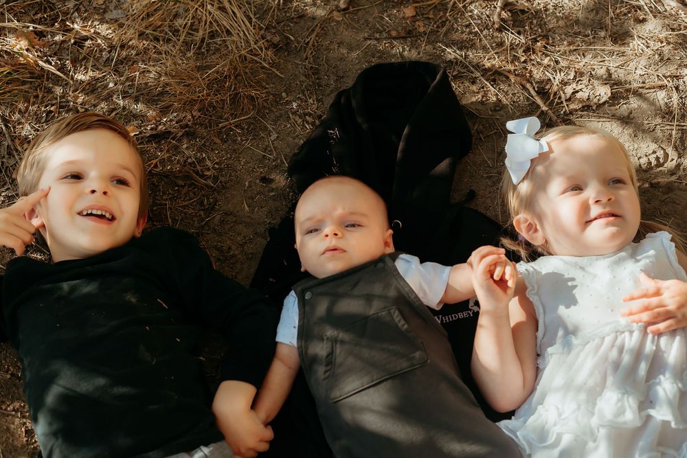 Three small children lie side by side on the ground wearing black and white outfits in natural lighting.