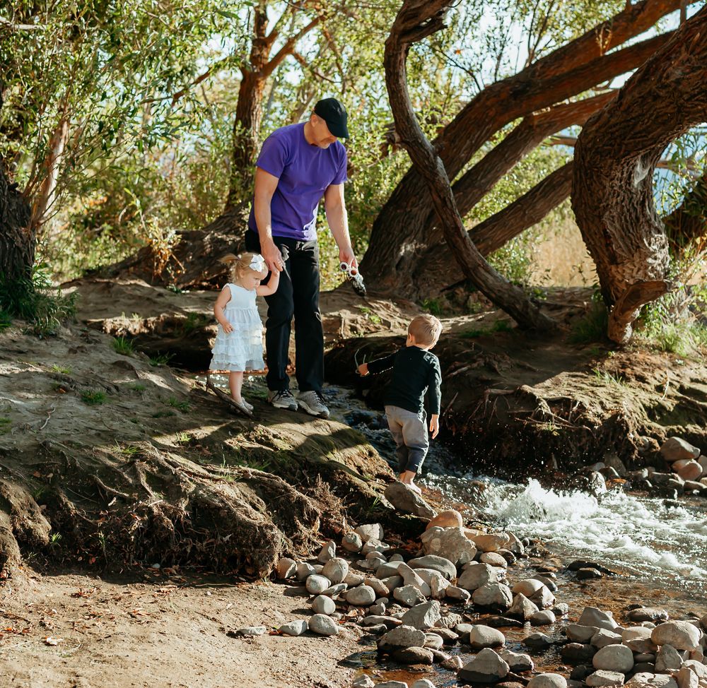 A figure in purple guides two young companions along a rocky stream bank under shady trees on a sunny afternoon.