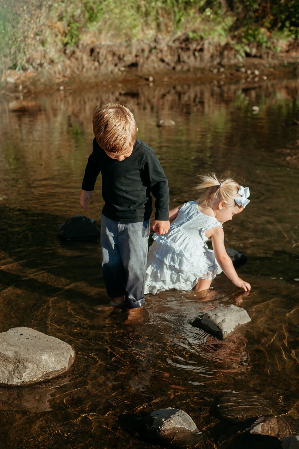 Two children explore a shallow creek with stepping stones on a sunny day, their reflections rippling in the clear water.