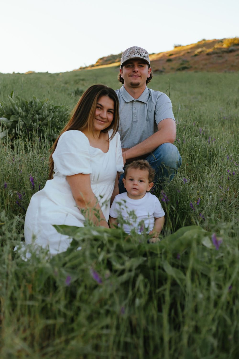 Family in white clothing sits together in a field of tall grass and wildflowers during golden hour.