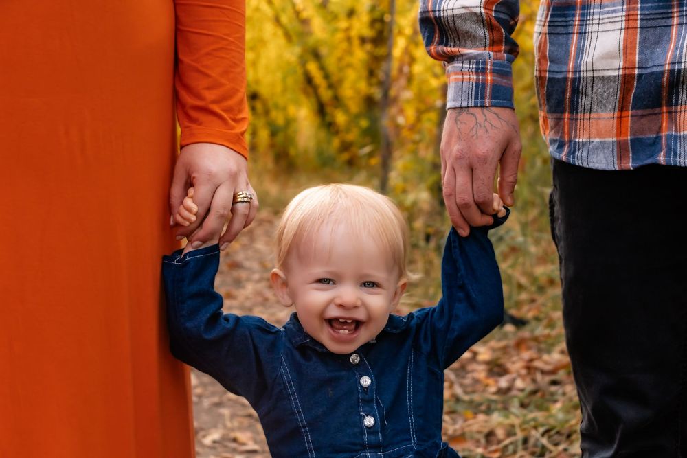 A happy child in a navy blue shirt holds hands with two adults while standing outdoors during autumn.