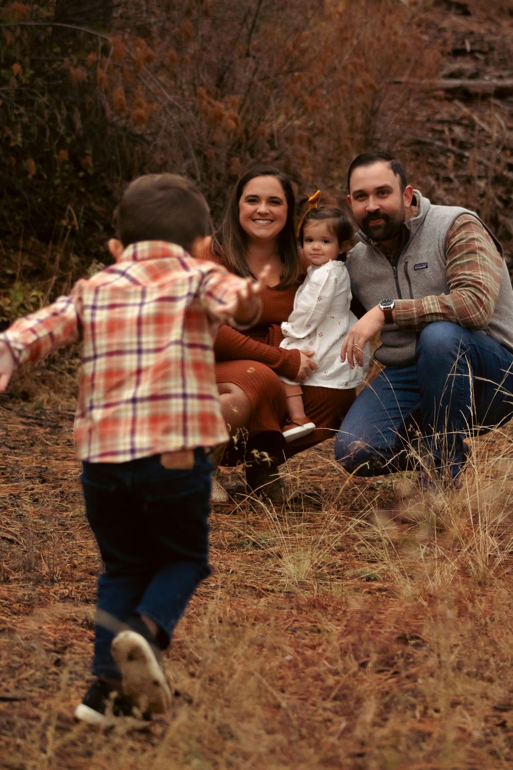 A family spends time together outdoors on an autumn day as a child in plaid runs towards them through dry grass.