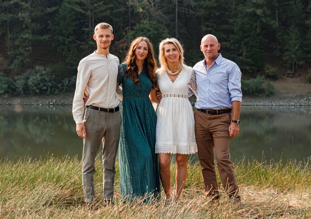 A group of four people stand together in casual attire by a tranquil lake with evergreen trees in the background.
