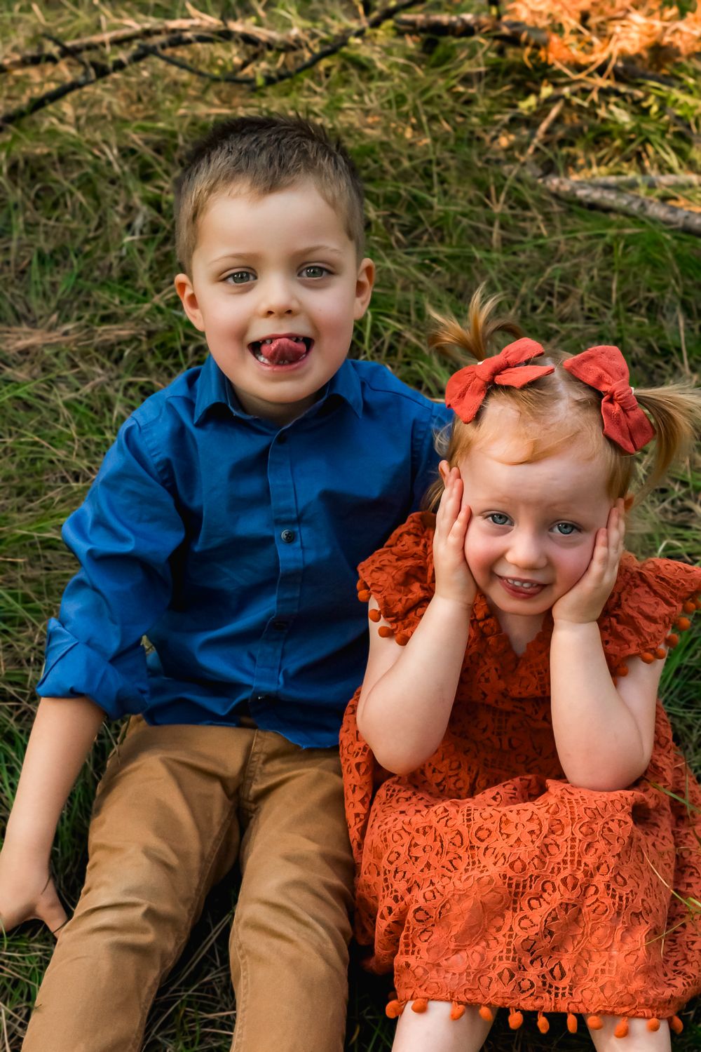 Two children in bright blue and orange outfits pose playfully on grass with autumn leaves in the background.