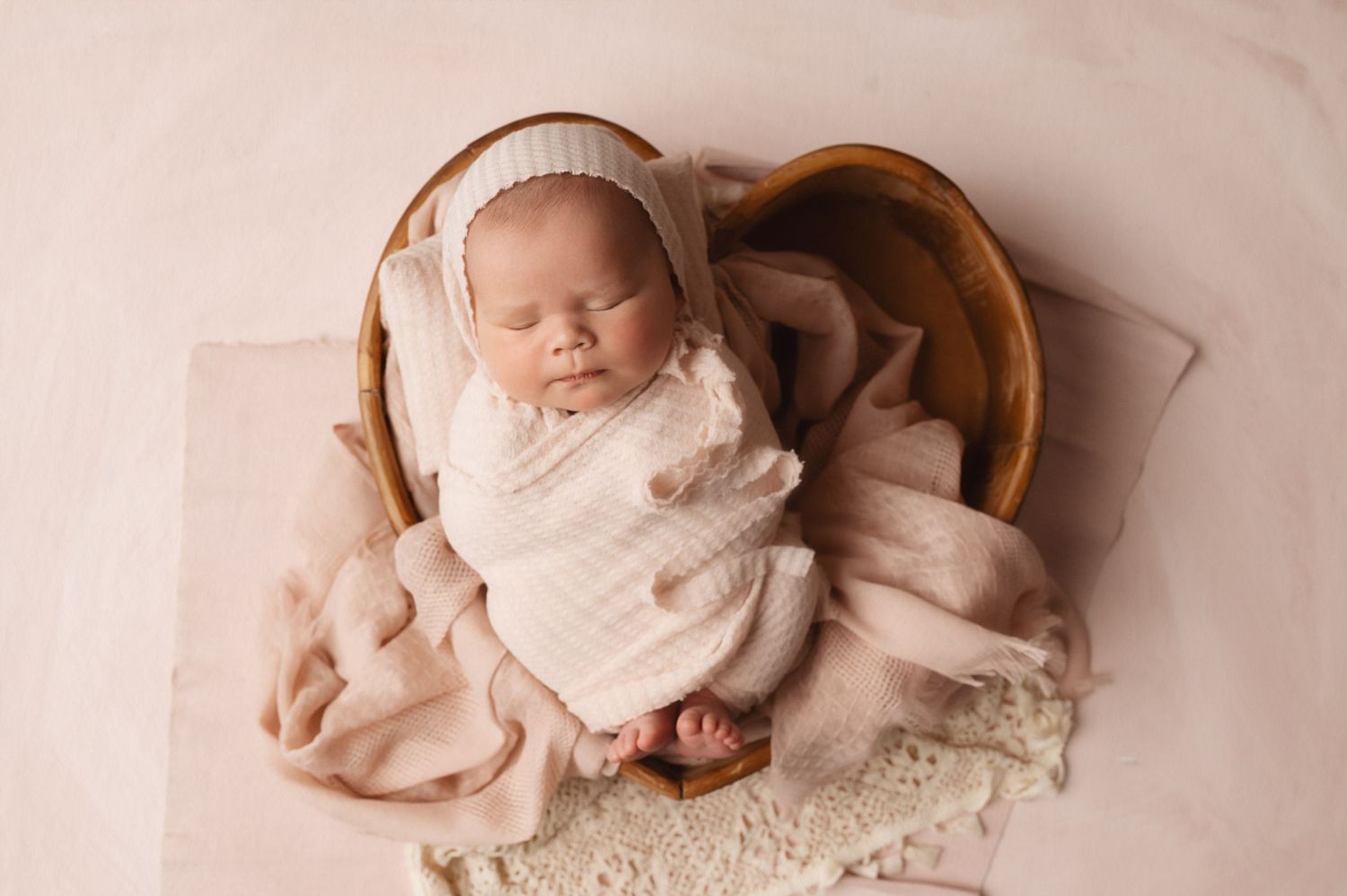 Peaceful newborn baby sleeping in cream wrap nestled in brown wooden bowl with lace details.