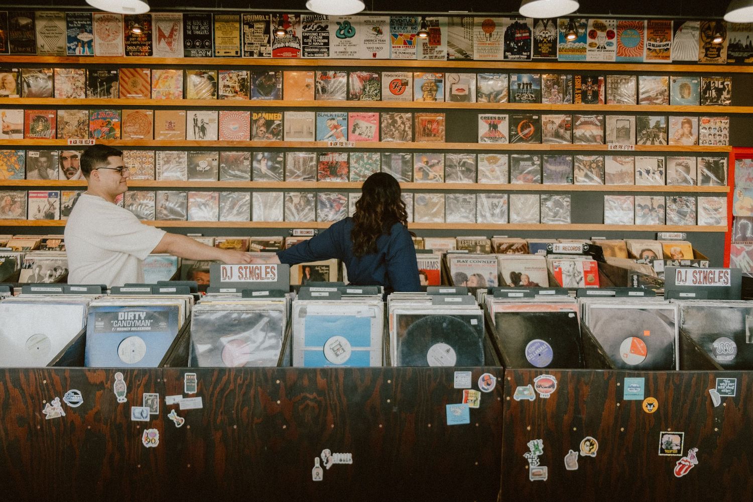 Two people browsing through vinyl records at a music store with wall of album covers.