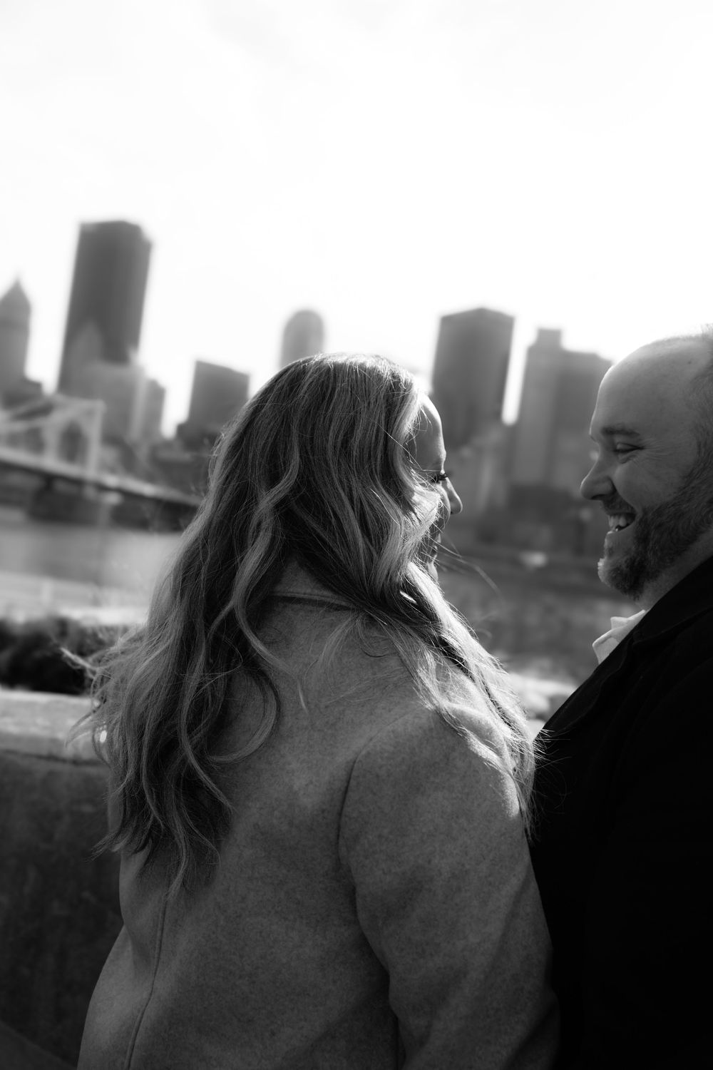A couple shares a romantic moment along the waterfront with a cityscape silhouette in black and white photography.