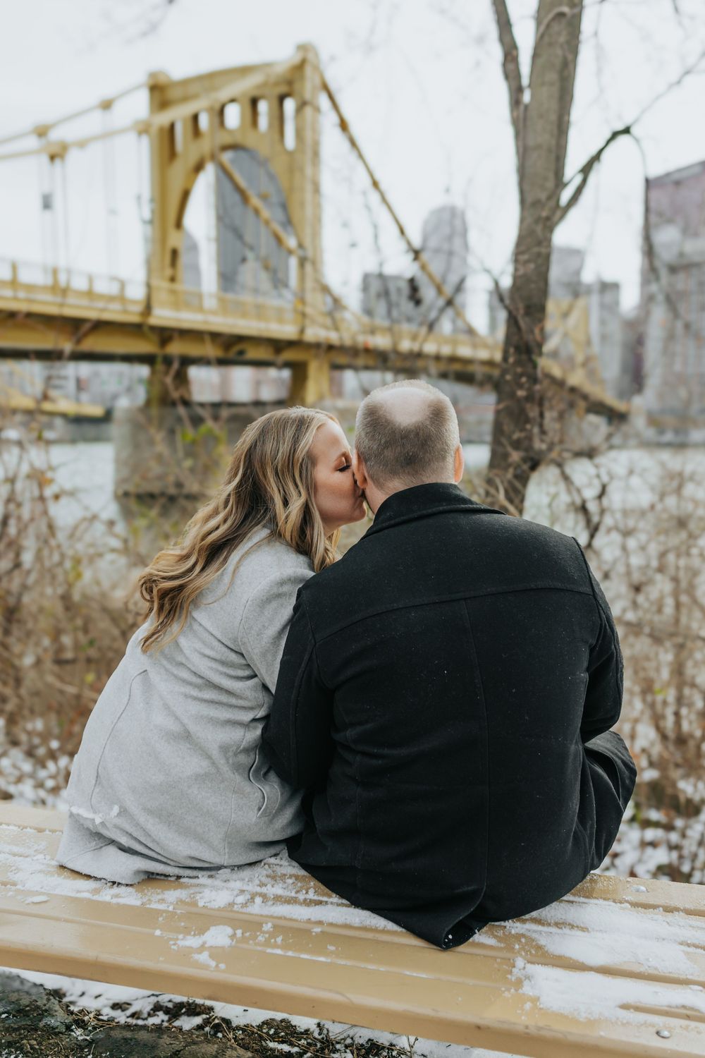 Couple sits together on bench near Pittsburgh's iconic yellow Roberto Clemente Bridge on winter afternoon.