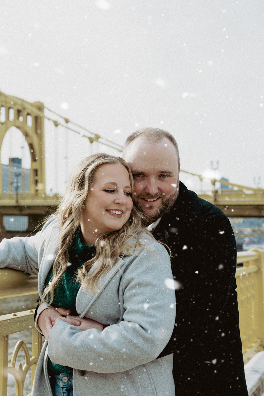 Happy couple embracing in front of iconic yellow bridge during snowfall in winter scene.
