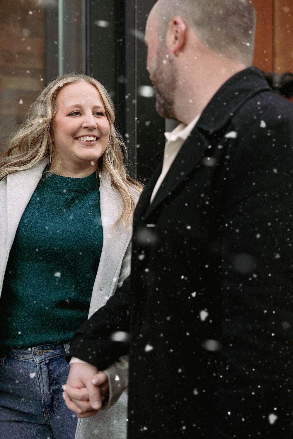 A couple shares a joyful moment together in the falling snow, with her wearing a green sweater and light coat.