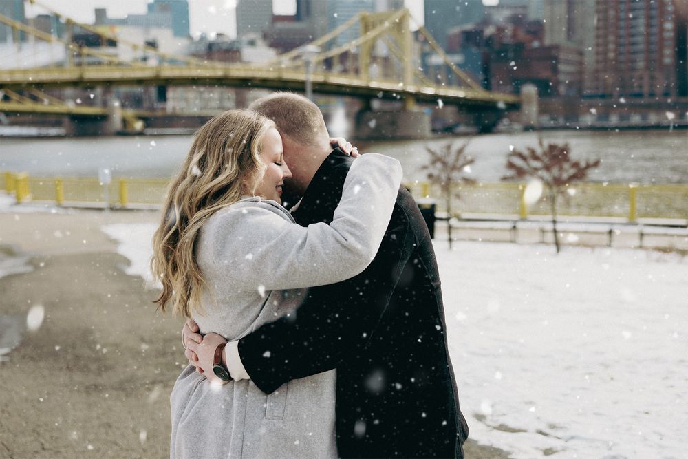 A couple shares a romantic kiss in the snow by a river with a yellow bridge visible in the background.