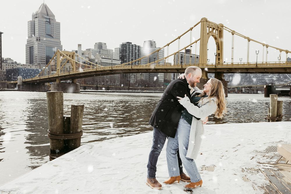 A romantic winter scene with couple embracing by the snowy riverside with Pittsburgh's Roberto Clemente Bridge in background.