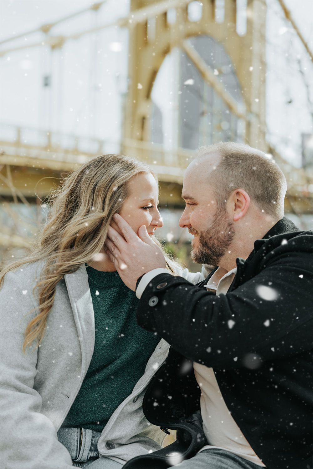 A romantic winter moment captured under a bridge as snow falls on a couple embracing in Pittsburgh.