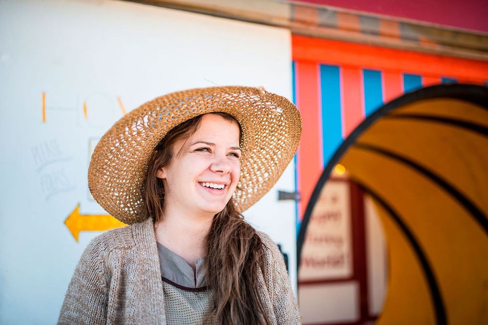 Someone wearing a straw sunhat and gray sweater smiles warmly against a colorful striped background.