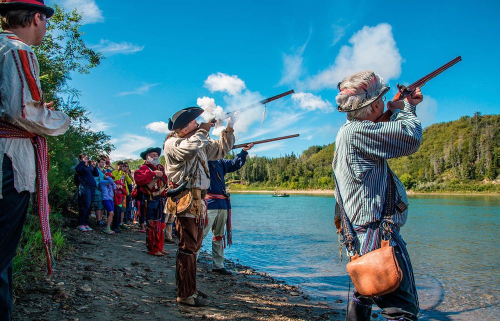 Historical reenactors in period costumes fire muskets along a riverbank during a living history demonstration.