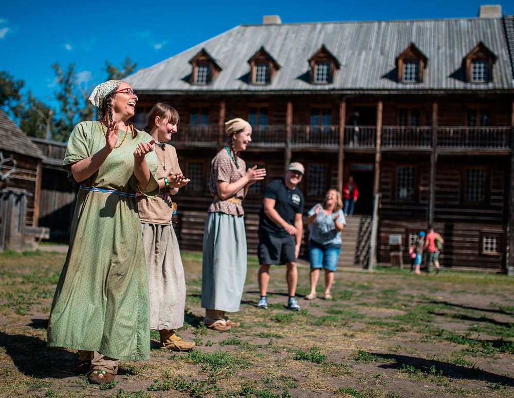 Period performers in historical costumes stand in front of a rustic log lodge with dormers at Fort Edmonton Park.