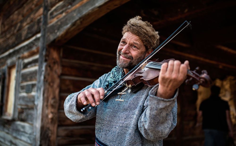 A cheerful musician plays violin outside a rustic log cabin on a bright day.
