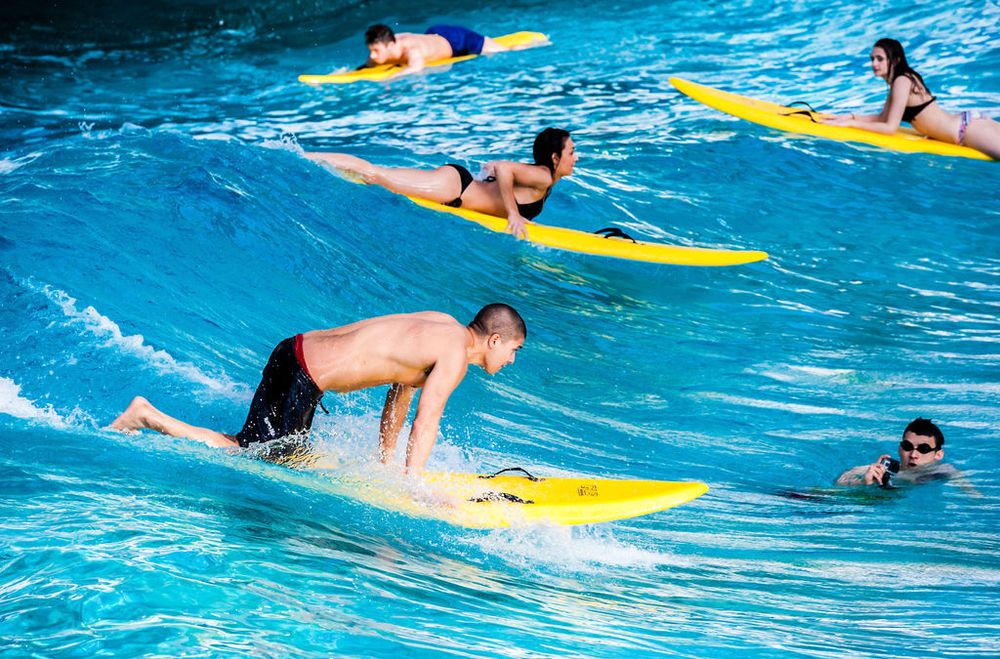 People practicing surfing techniques on yellow boards in a bright blue swimming pool.