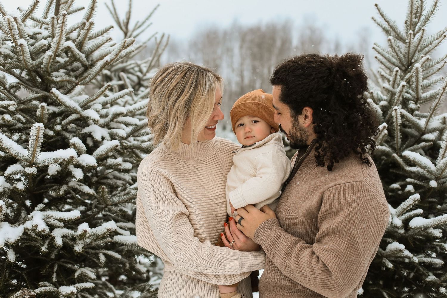 Family wearing cozy sweaters poses together among snow-covered evergreen trees at a Christmas tree farm.