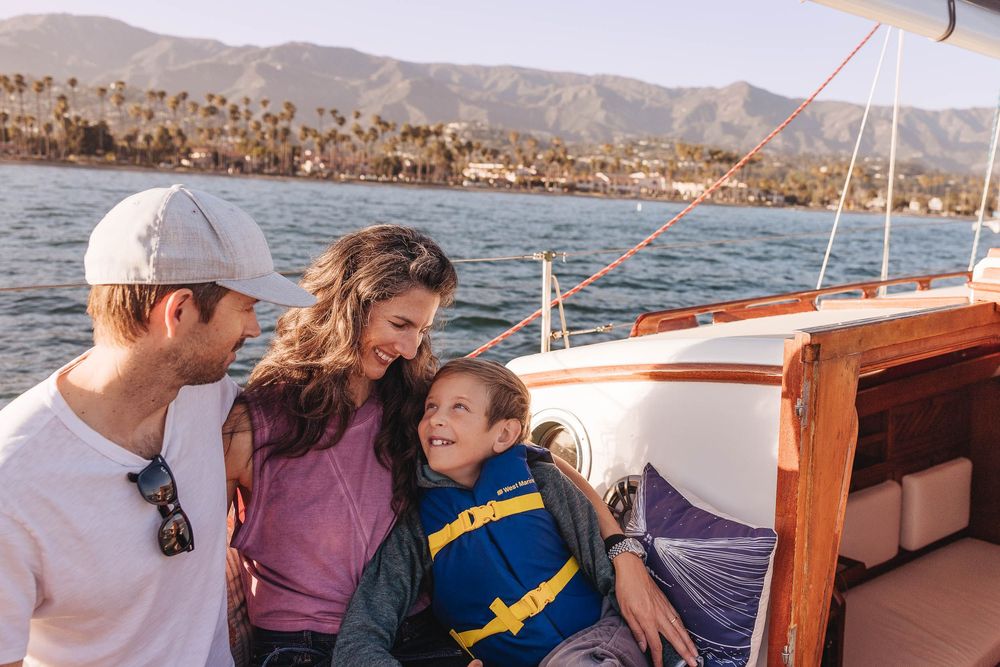 A group enjoys a sunny day on a sailboat with mountains and coastline visible in the background.