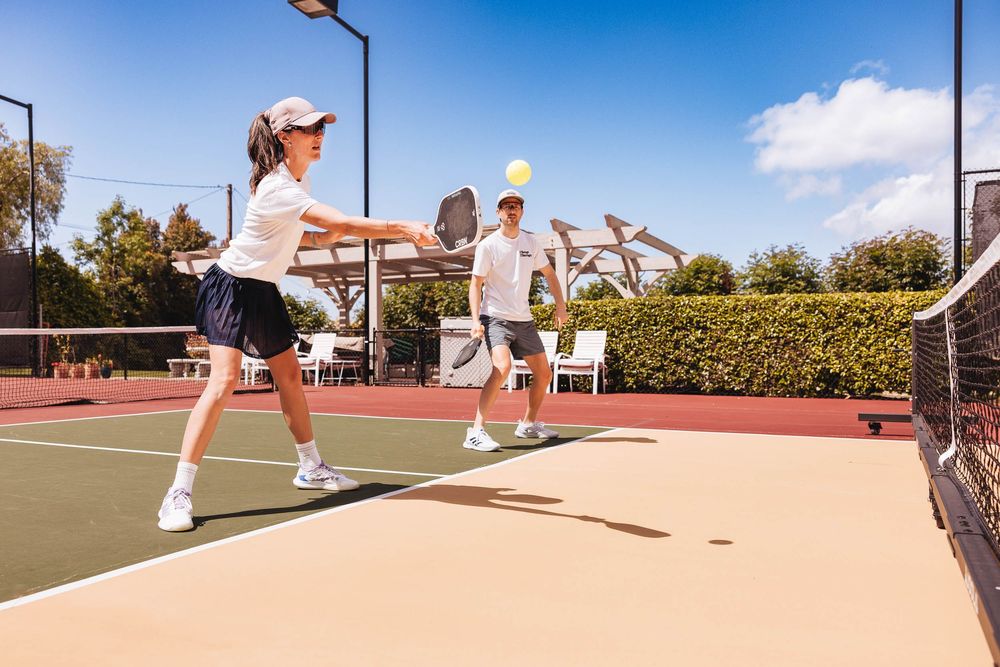 Two individuals playing tennis on an outdoor court with manicured hedges and a sunny blue sky in the background.