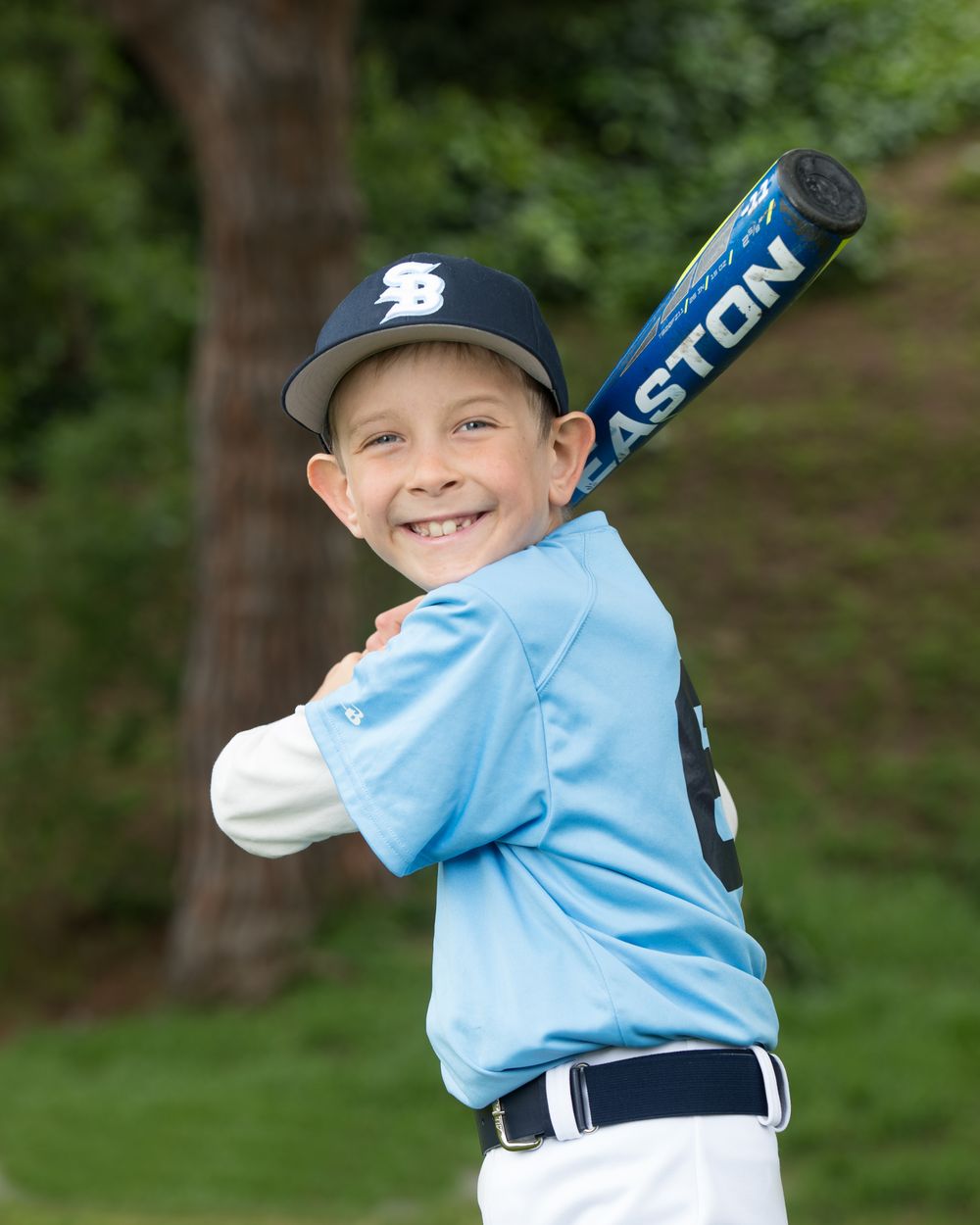 Young baseball player in light blue jersey and navy cap poses with Easton bat during team photo day in outdoor setting.
