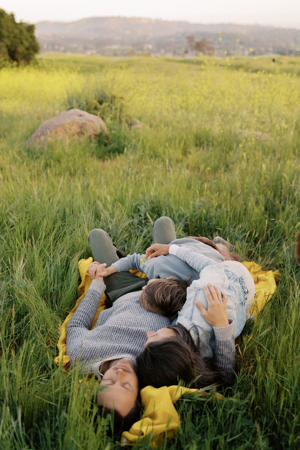 A couple rests peacefully together in tall grass during golden hour, wearing gray sweaters and yellow accessories.