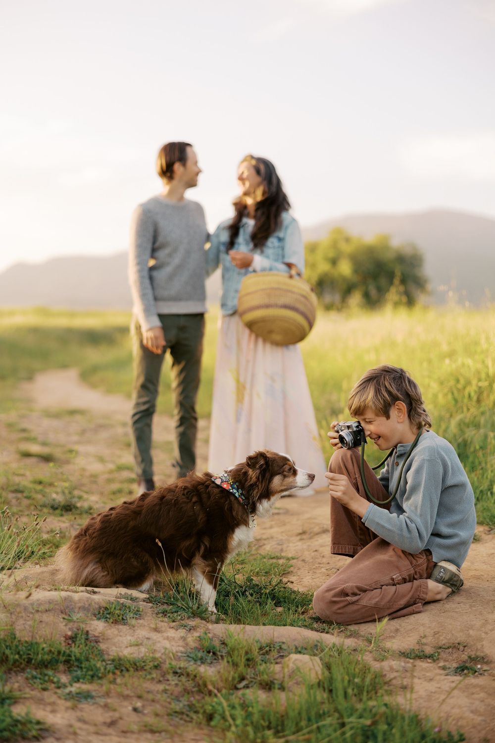 Photographer captures candid moments in a field at golden hour while a brown dog rests nearby on a dirt path.