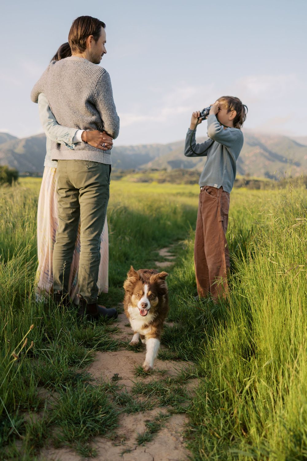 Two hikers and a collie dog explore a scenic mountain trail surrounded by tall grass and rolling hills.