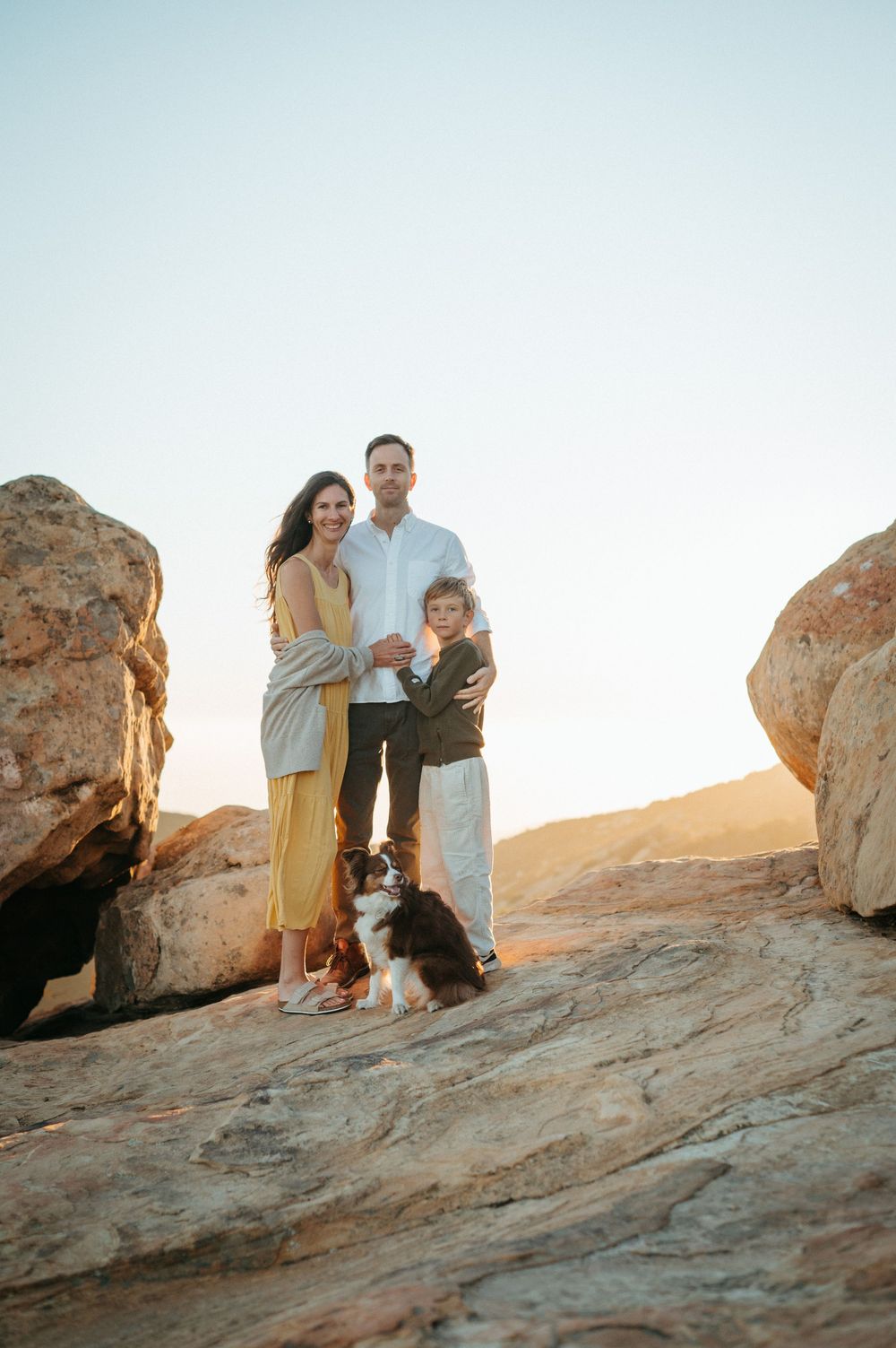 A family and their dog pose together on red rocks during sunset in a desert landscape.