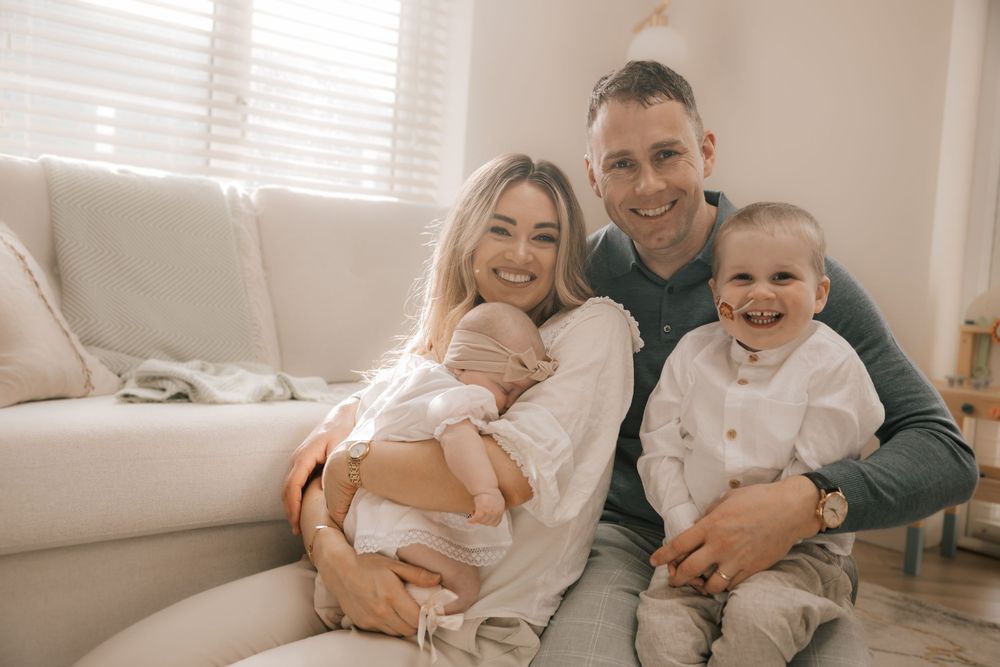 A family sits together on a white couch in a bright room with sunlight streaming through window blinds.