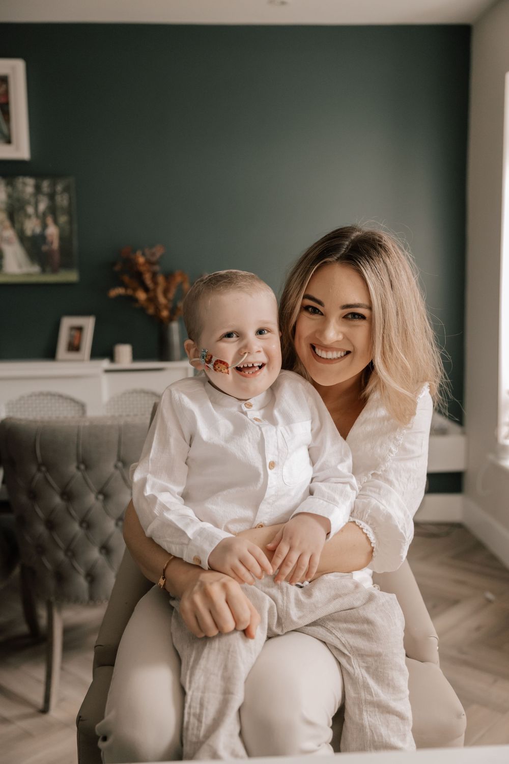 A child and adult wearing matching white outfits sit together on a gray tufted chair in a room with dark green walls.