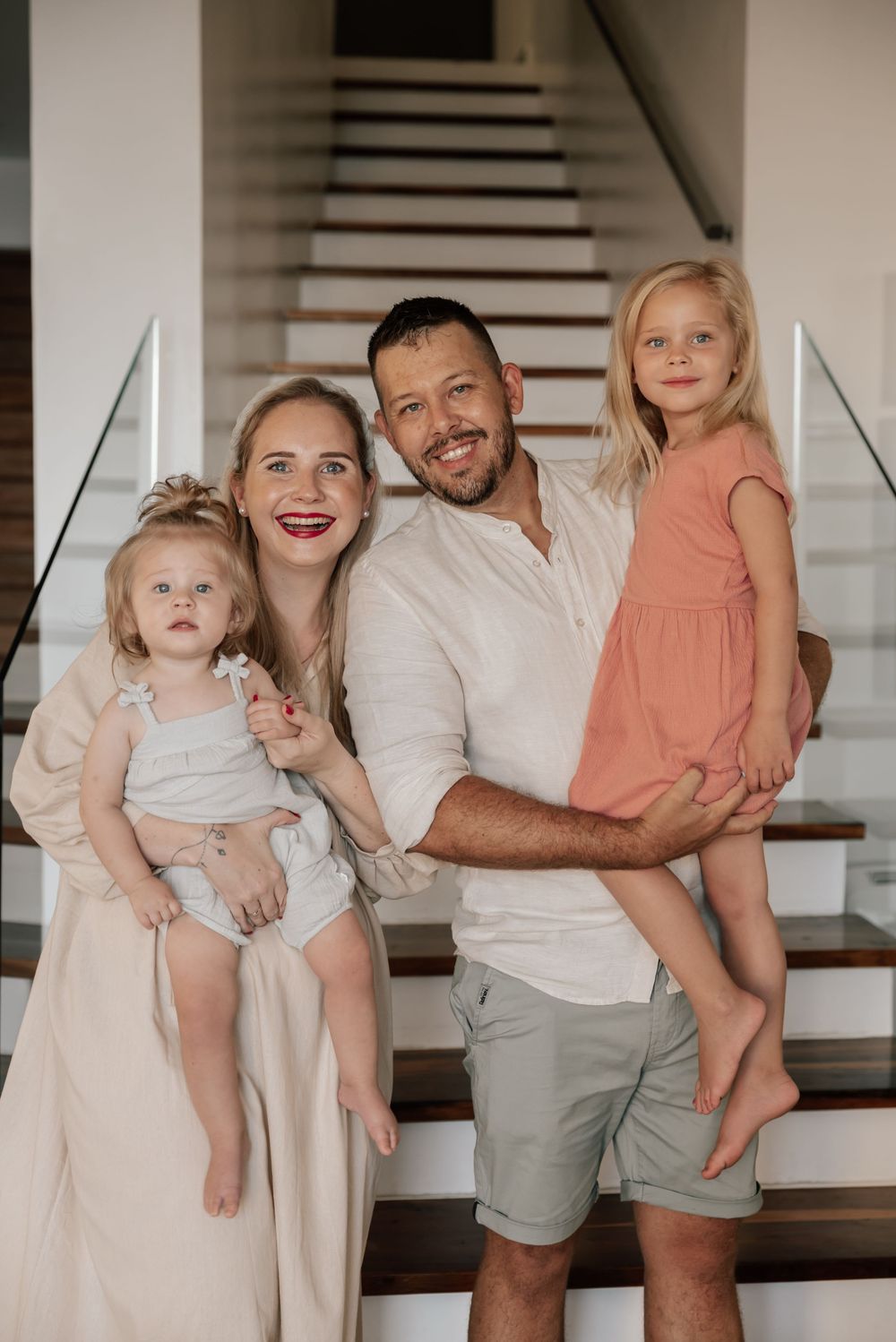 A family in coordinated neutral outfits poses together on a white staircase in their home.