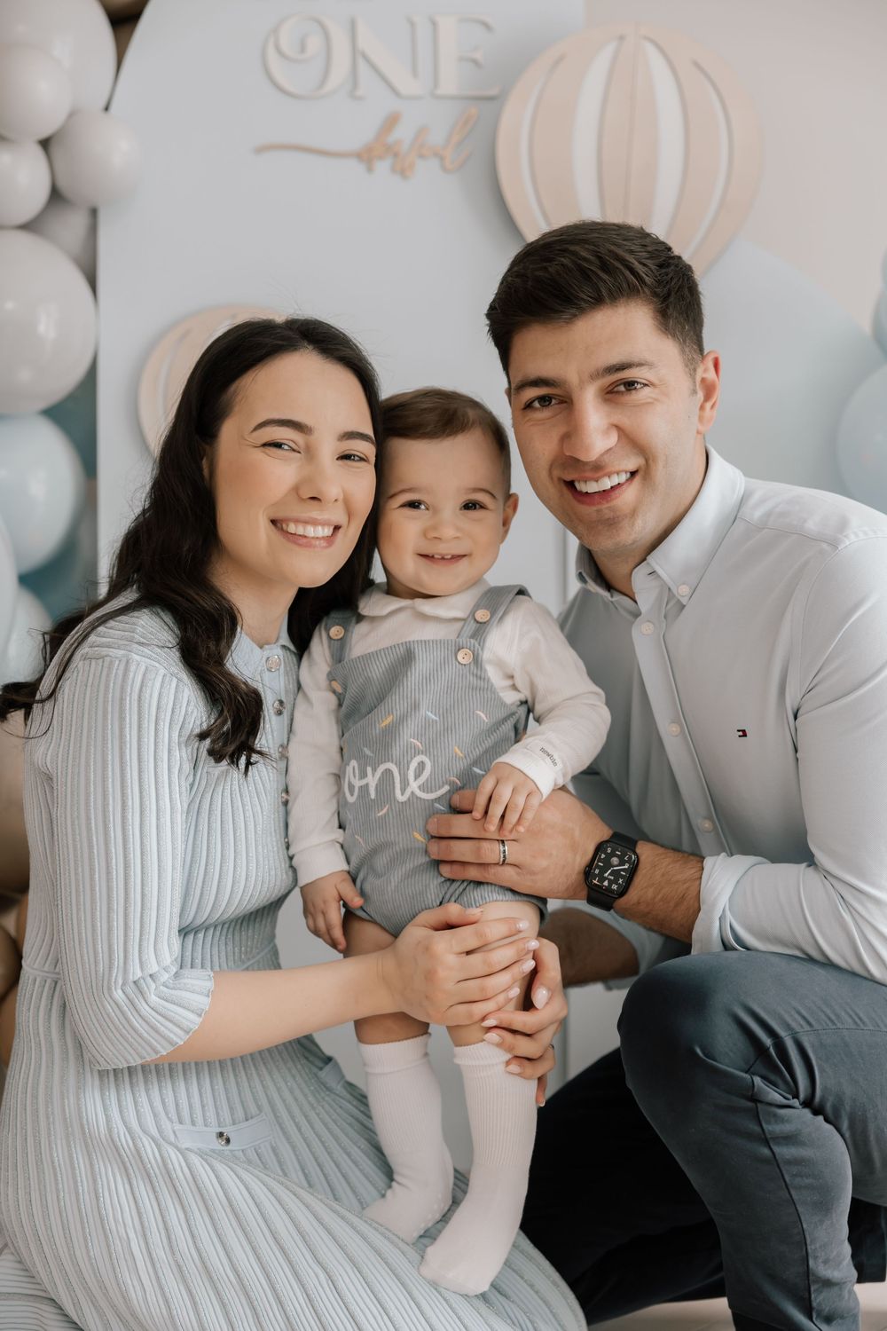 A family poses together against a white balloon backdrop with a 'ONE' decoration for a first birthday celebration.
