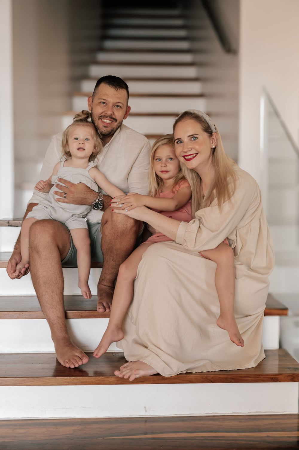 Family sitting together on white stairs wearing neutral beige tones in a bright interior setting.
