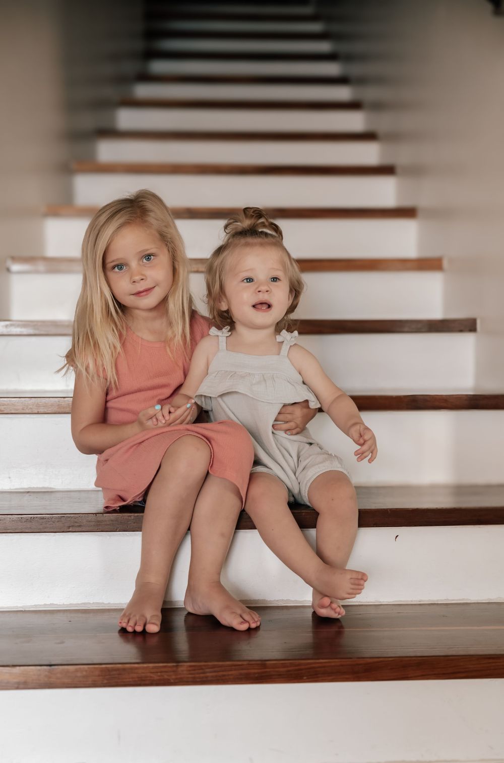 Two young children sit together on white wooden stairs wearing casual summer outfits and bare feet.