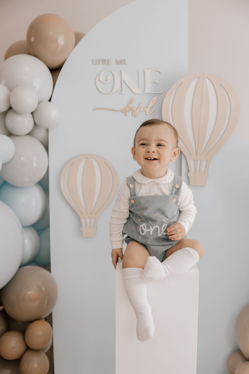 First birthday celebration setup with neutral balloons, hot air balloon decorations and child seated in white outfit against white backdrop.