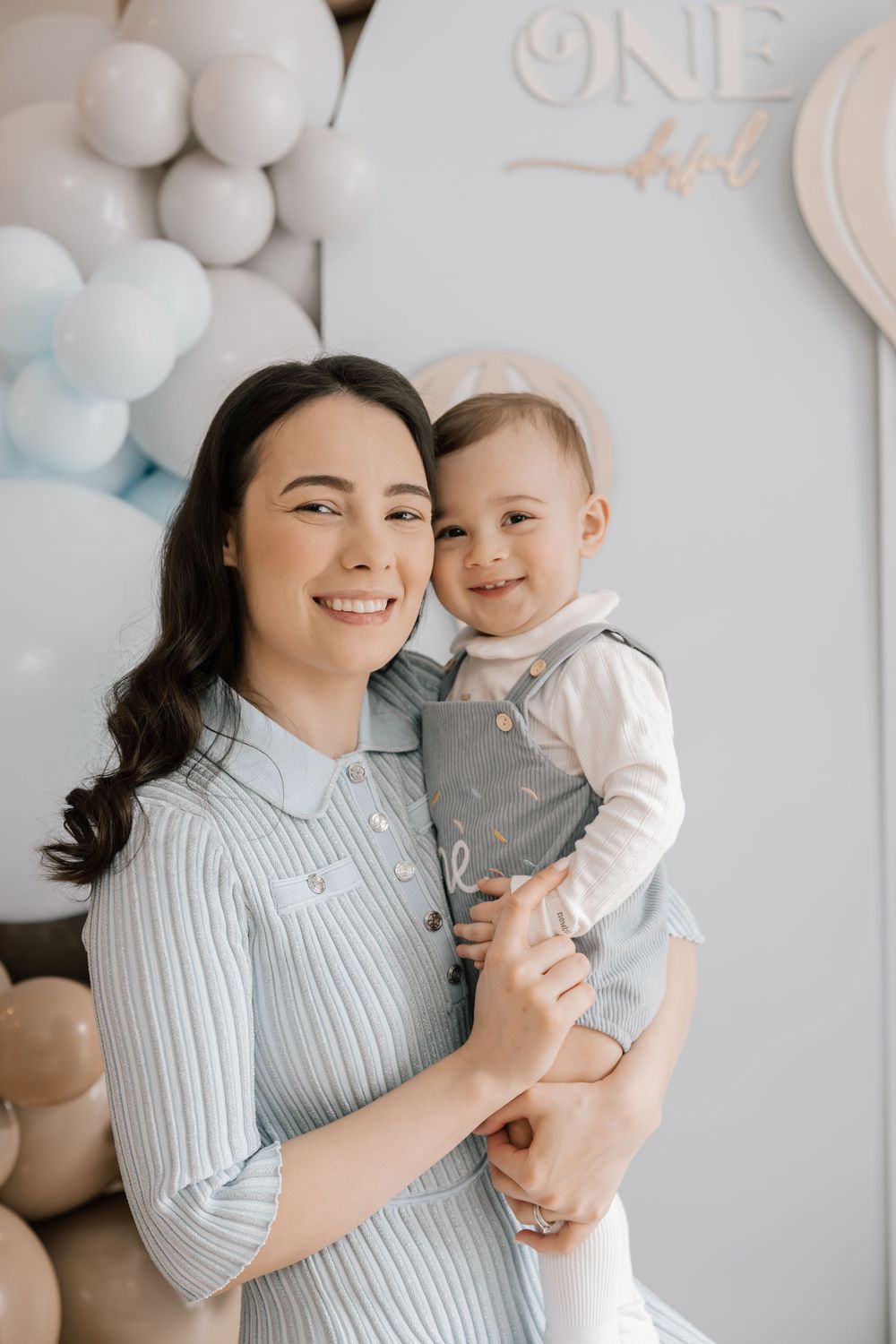 A parent and child pose together against light blue and white balloons at a first birthday celebration party.