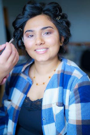 Someone wearing a blue plaid flannel shirt and gold necklace smiles while having makeup applied.