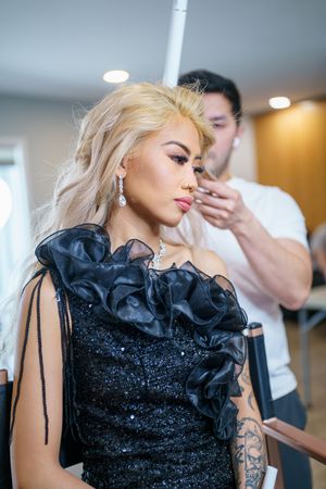 Stylist works on arranging blonde hair while model wears elegant black sequined gown with ruffled neckline detail.